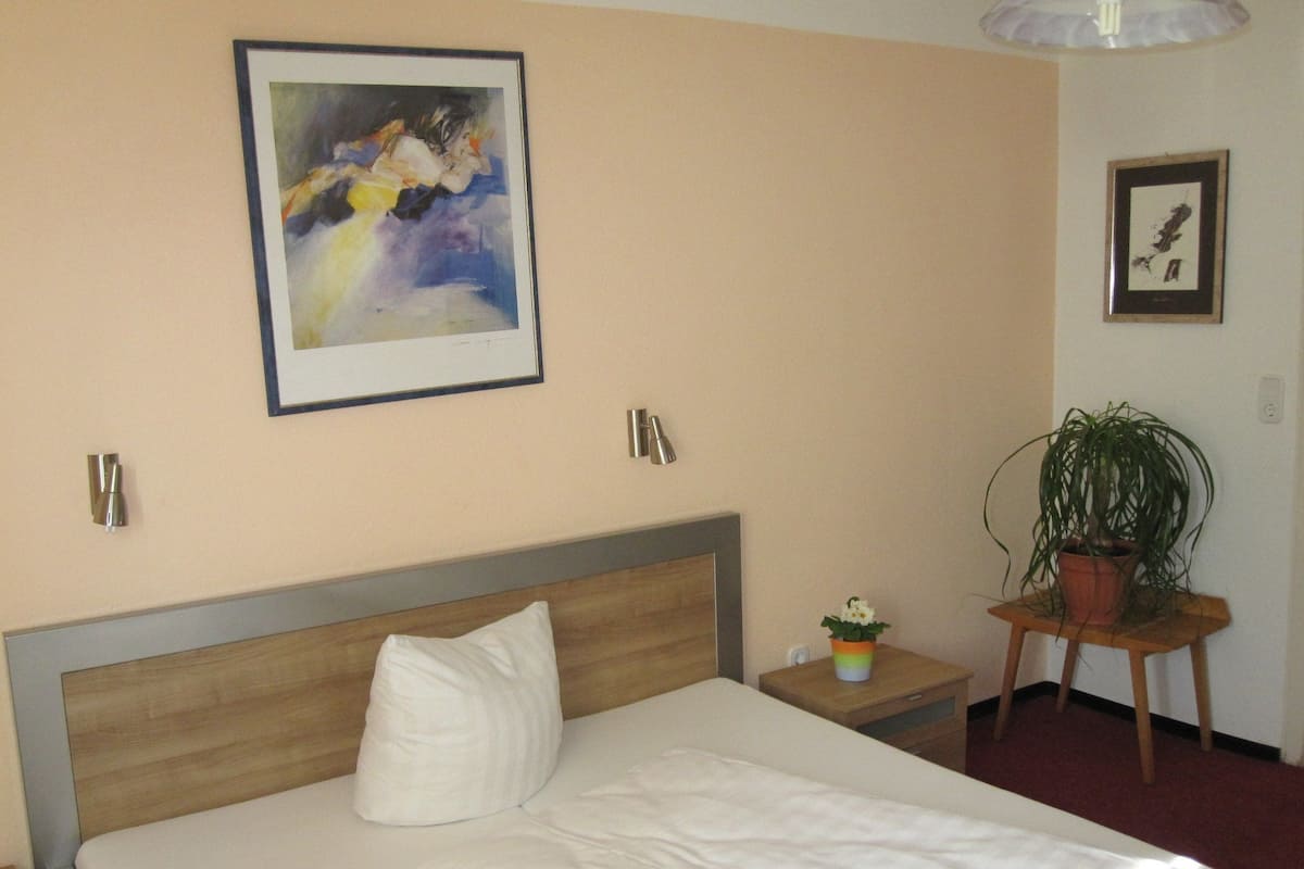 A hotel room with a bed, a nightstand, a potted plant, and two framed artworks on the walls.