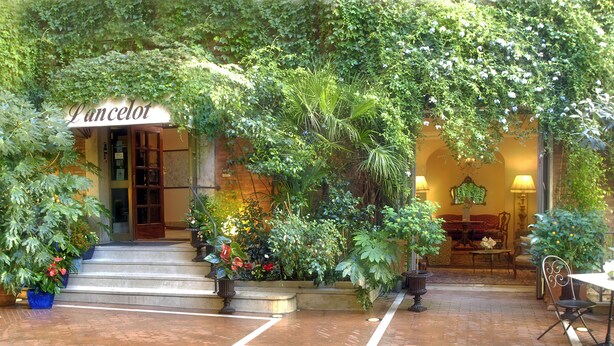 Entrada de hotel con un letrero que dice "Lancelot", rodeada de abundante vegetación y plantas en macetas.
