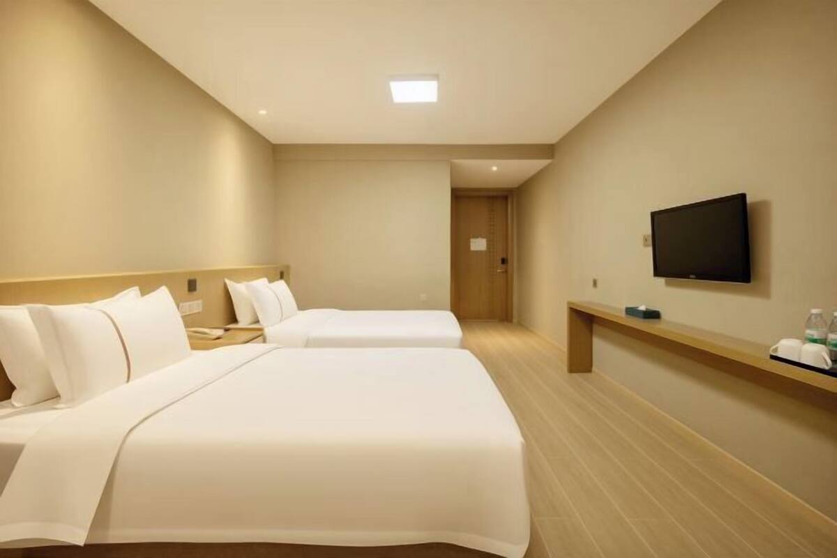 A hotel room with two beds, a TV on the wall, a bedside table, and a wooden bench.