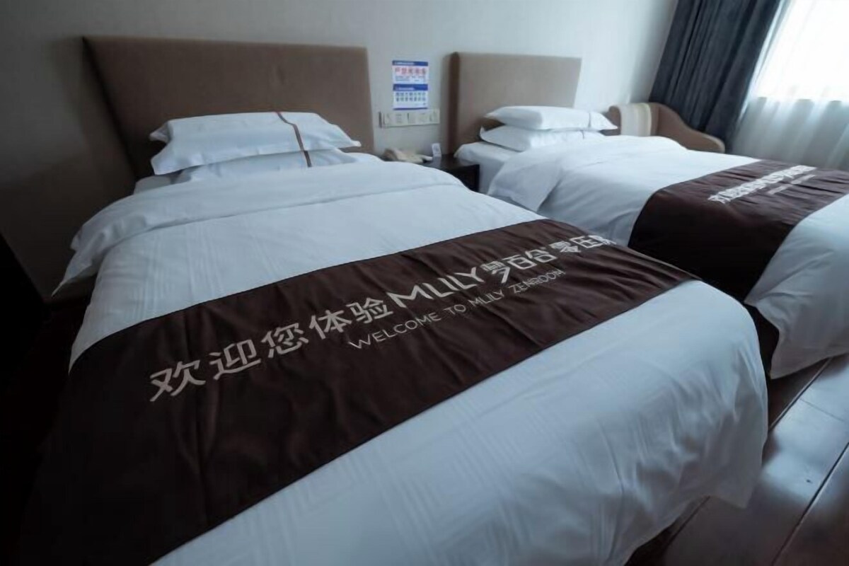 A hotel room with two beds, each with a brown bedspread and white linens. There is a wall-mounted clock and a sign with text in a foreign language.