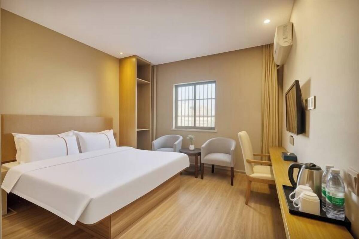 A hotel room with a bed, two chairs, a small table with a kettle and cups, a TV, and a window with curtains.