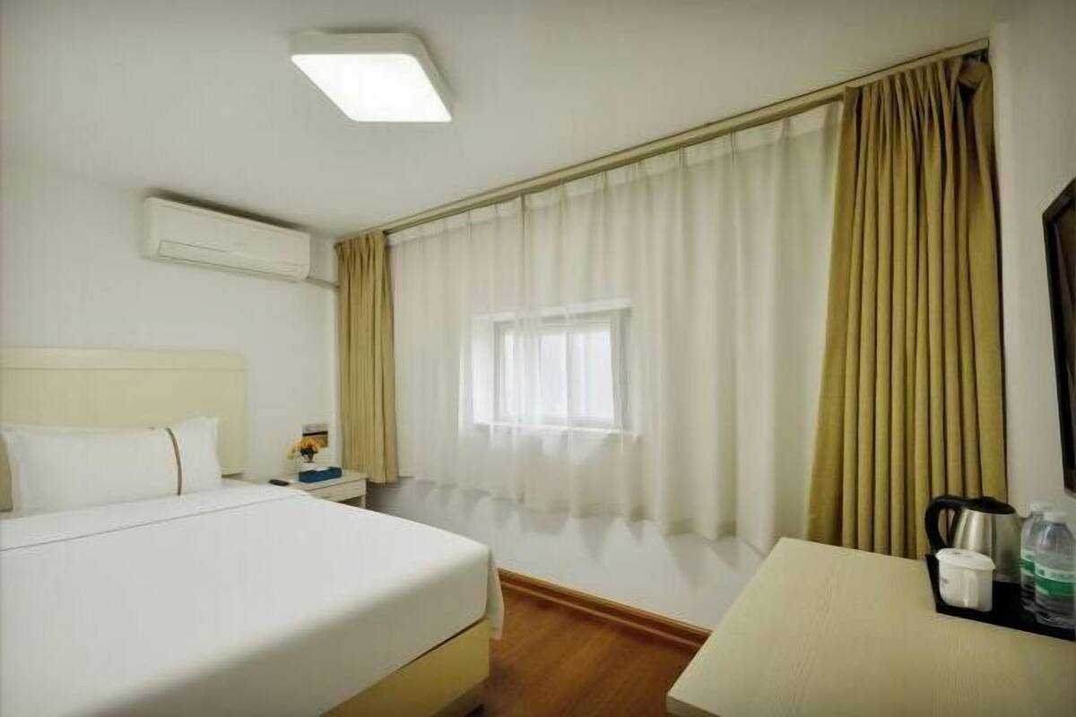 A hotel room with a bed, bedside table, a small table with a kettle and cups, a window with curtains, and an air conditioning unit.