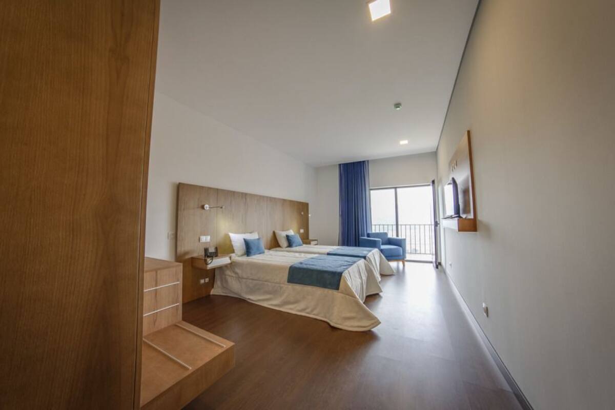 Ein modernes Hotelzimmer mit einem hölzernen Kopfteil, zwei Betten, einem Schreibtisch und einem großen Fenster mit Vorhängen.