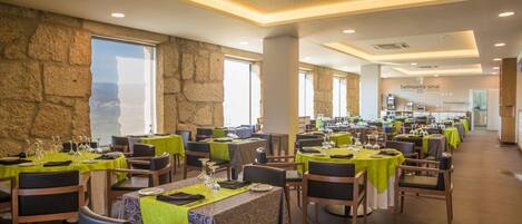 Un restaurant doté de nombreuses tables, chacune recouverte d’une nappe verte et agrémentée de verres à vin, d’assiettes et de couverts. La salle est caractérisée par des murs en pierre et de grandes fenêtres offrant une vue sur la mer.
