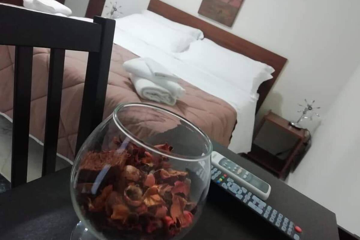 A hotel room with a bed, a bedside table with a remote control, and a glass bowl of snacks on the table.