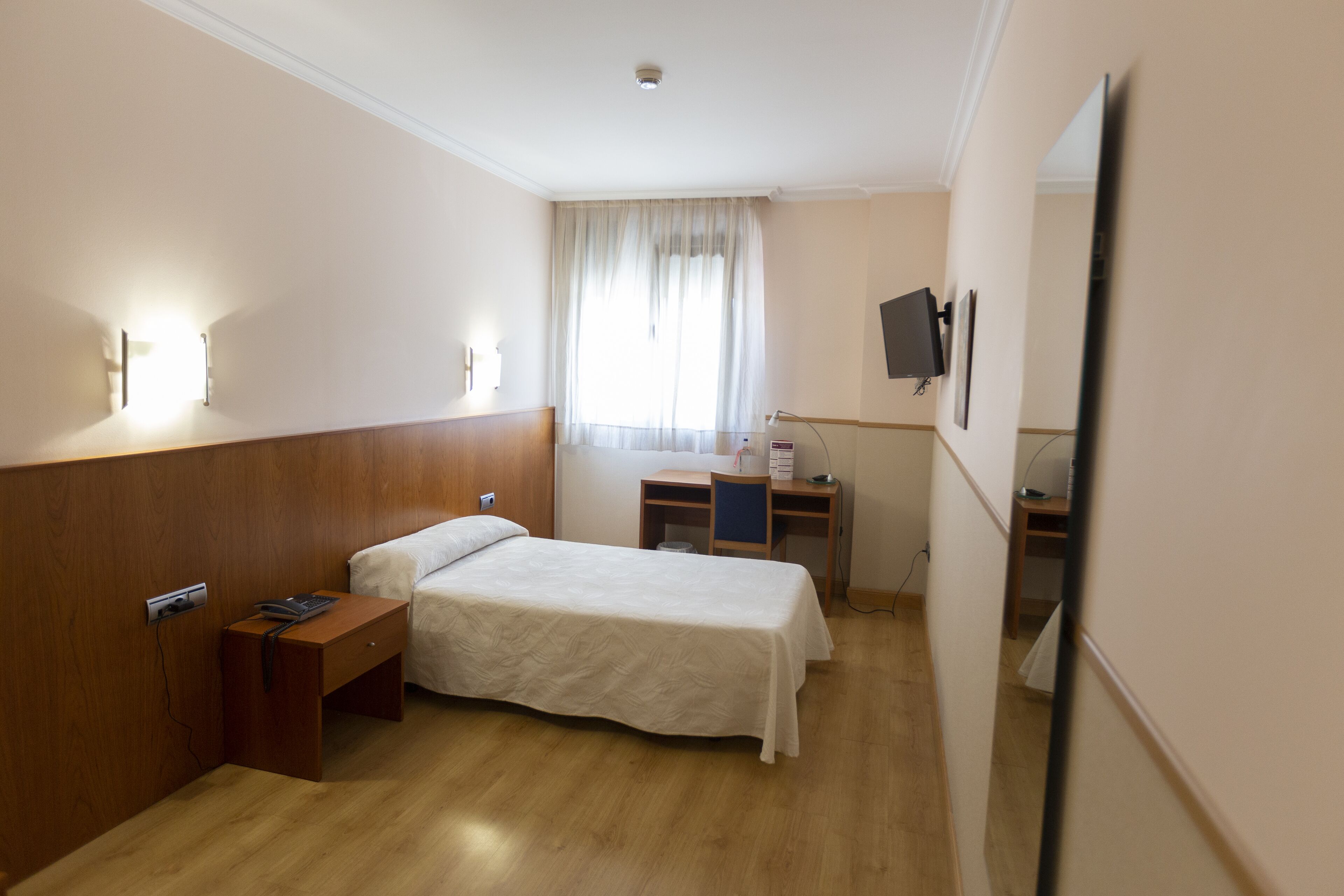 Habitación de hotel con cabecera de madera, una cama individual, un escritorio con silla, un televisor de pantalla plana montado en la pared y una ventana con cortinas.