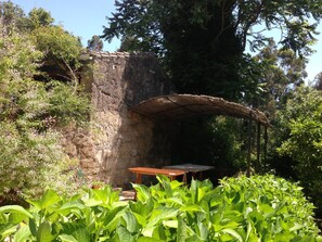 A rustic stone structure with a wooden bench and table under a thatched roof, surrounded by lush greenery. - Casa O Fabal (Sanxenxo)