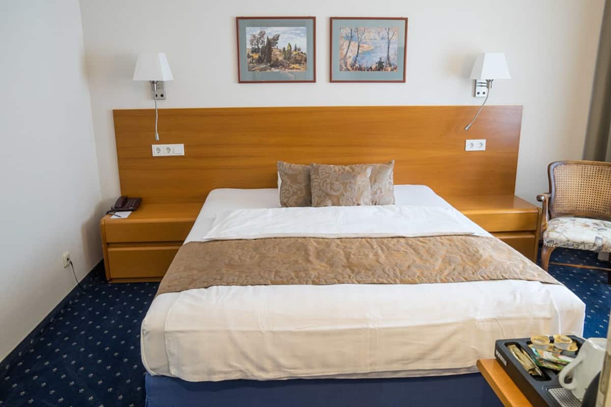 A hotel room with a large bed, two bedside lamps, a wooden headboard, a small bedside table with a telephone, and two framed paintings on the wall.