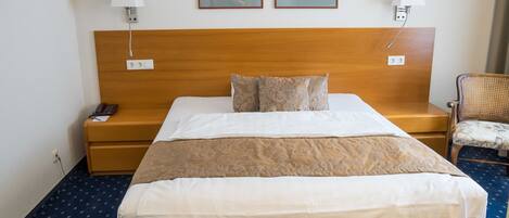 Habitación de hotel con una cama grande, dos lámparas de noche, un cabecero de madera, una mesita de noche con teléfono, y dos cuadros enmarcados en la pared.