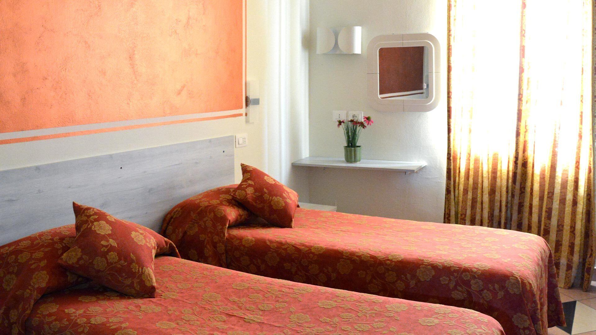 A hotel room with two beds, each with floral-patterned bedding. There is a small window with a mirror above the bed, and a vase with flowers on a nightstand.