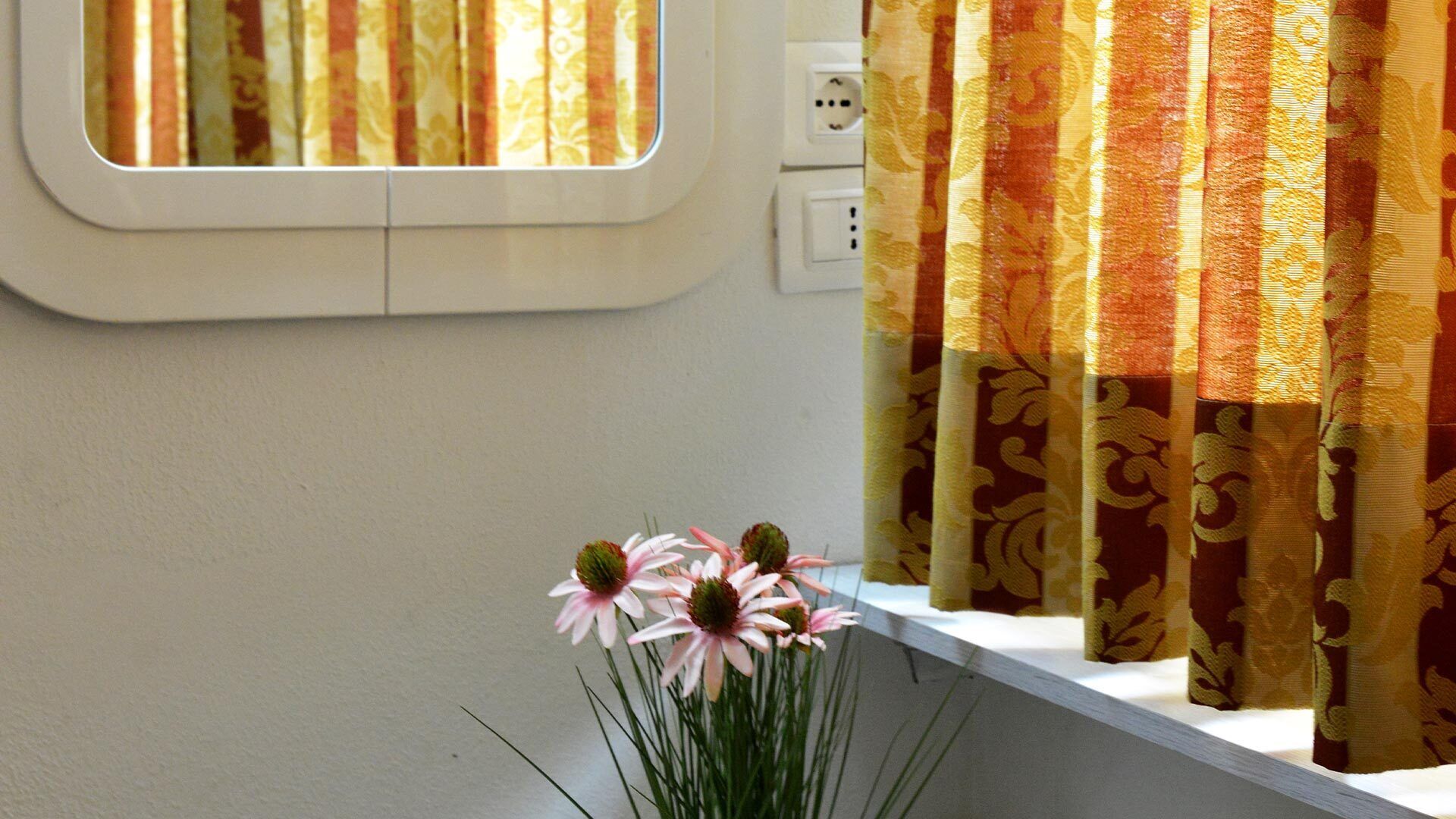 A window with curtains, a power outlet, and a vase with flowers on a windowsill.