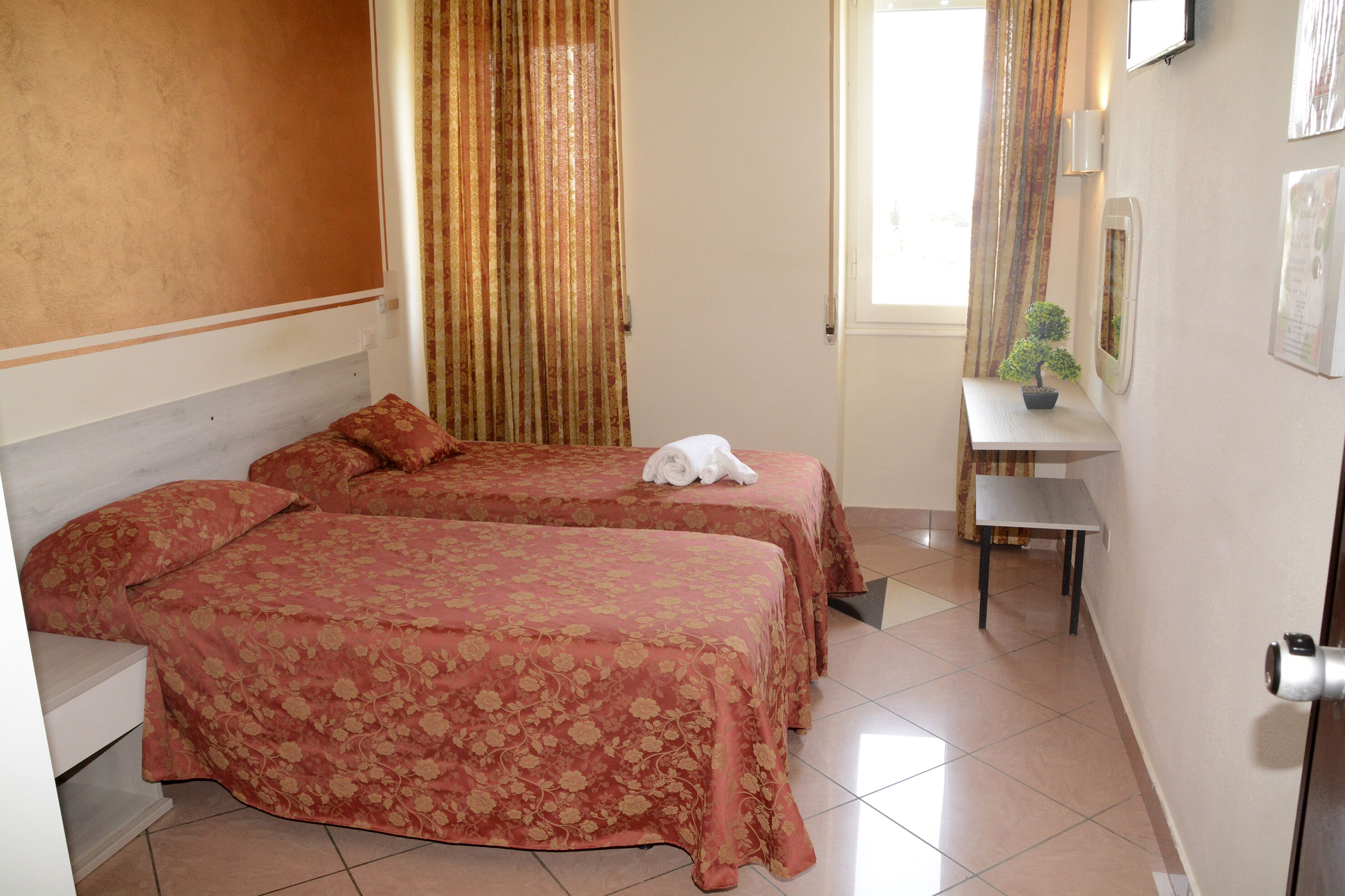 A hotel room with two beds, a small desk, a potted plant, and a window with curtains.