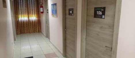 Egy szállodai folyosó, csempézett padlóval, fa ajtókkal és egy tűzoltó készülékkel.