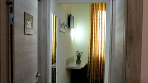 Un pasillo con una puerta de madera identificada con el número 203, piso de baldosas, una mesita con una planta en maceta y una ventana con cortinas.