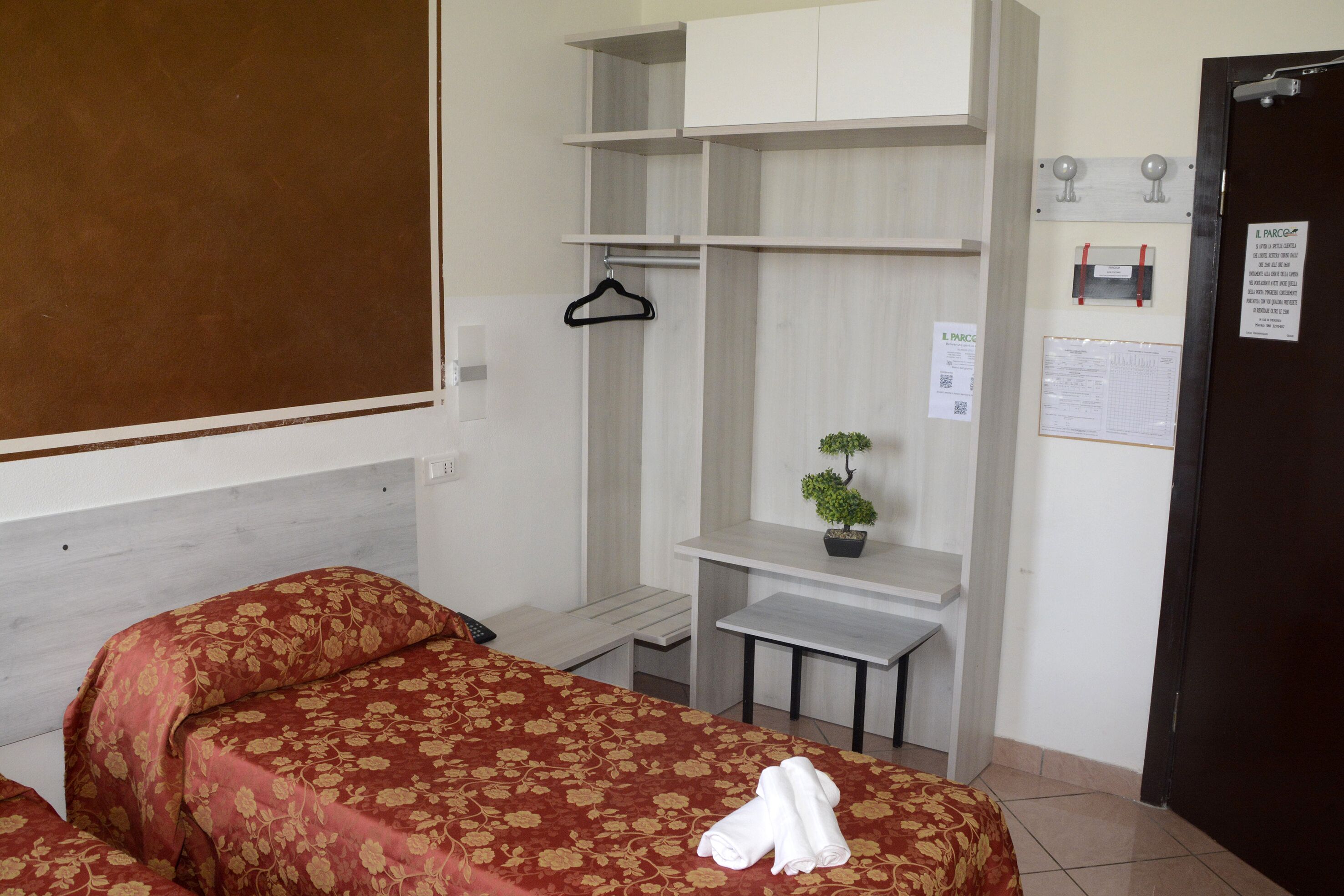 A hotel room with a bed, a desk, a small table, a hanging coat rack, and a potted plant.