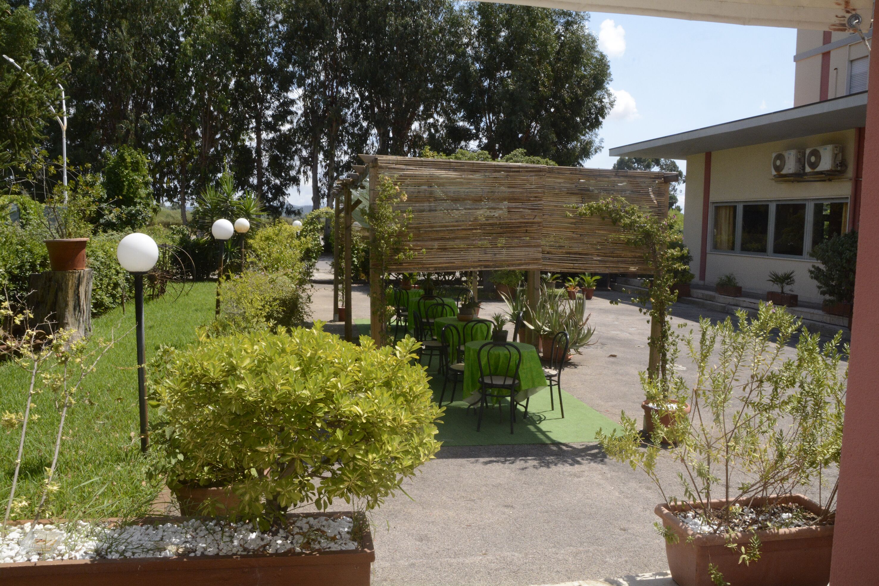 An outdoor patio area with tables and chairs, surrounded by potted plants and a wooden pergola.
