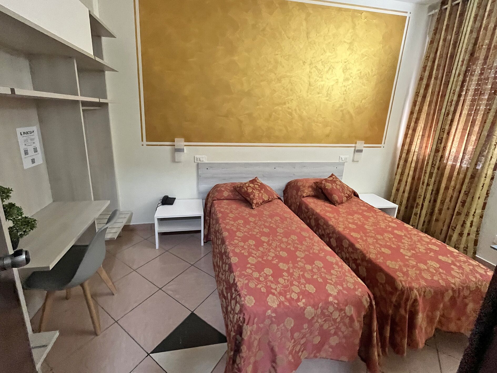 A hotel room with two single beds, a desk, a chair, a small table, and a large window with curtains.