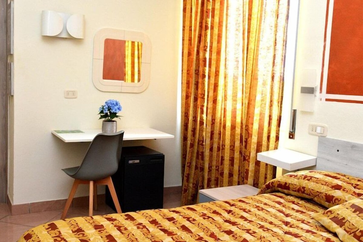 A hotel room with a bed, a desk with a chair, a small refrigerator, a bedside table, and a curtain.