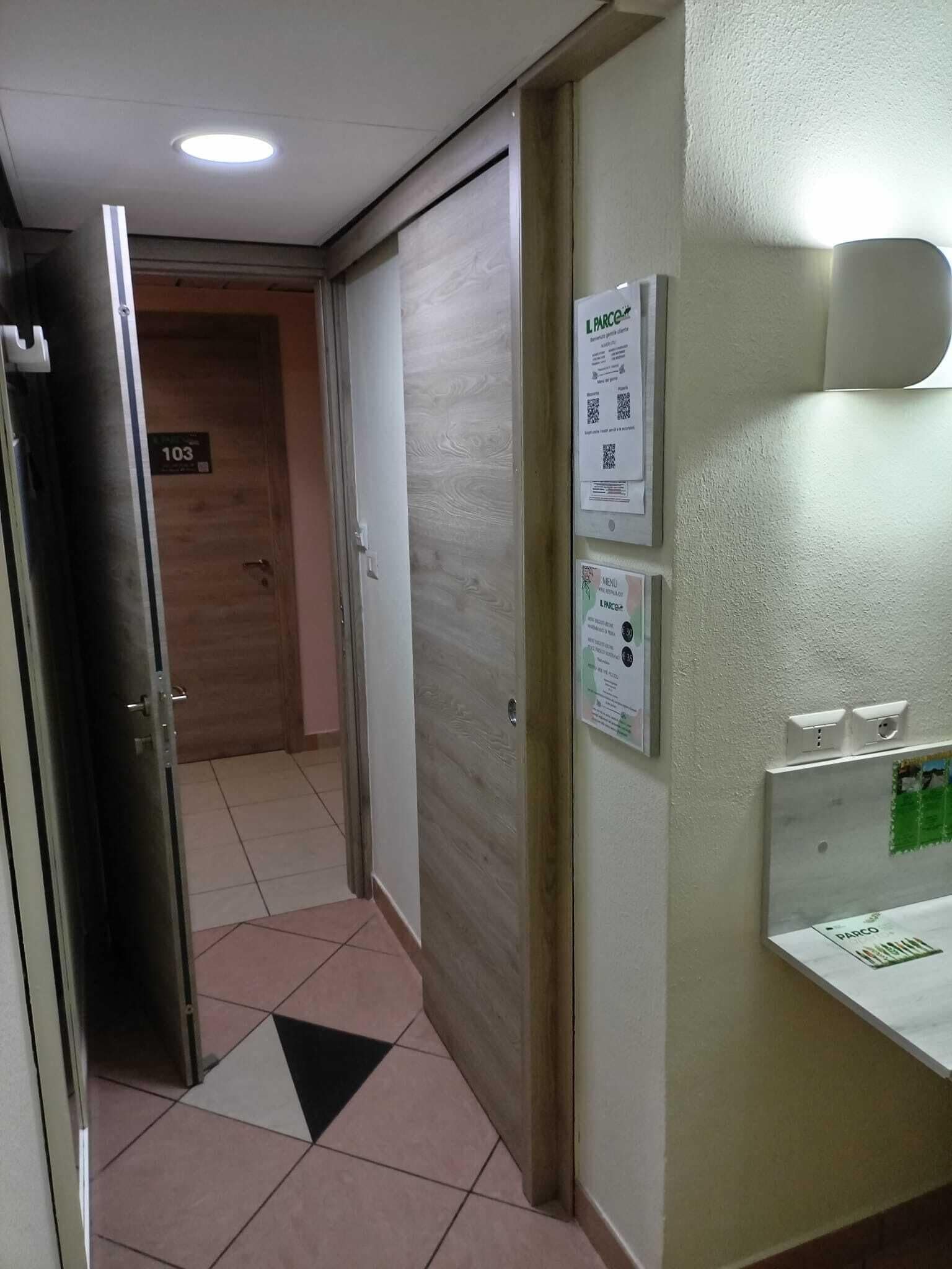 Ein Flur mit einer Tür, auf der „103“ steht, die zu einem Raum führt. An der Wand befinden sich Schilder, die ein Restaurant namens „L PARCO“ mit einer Speisekarte und einem QR-Code bewerben.