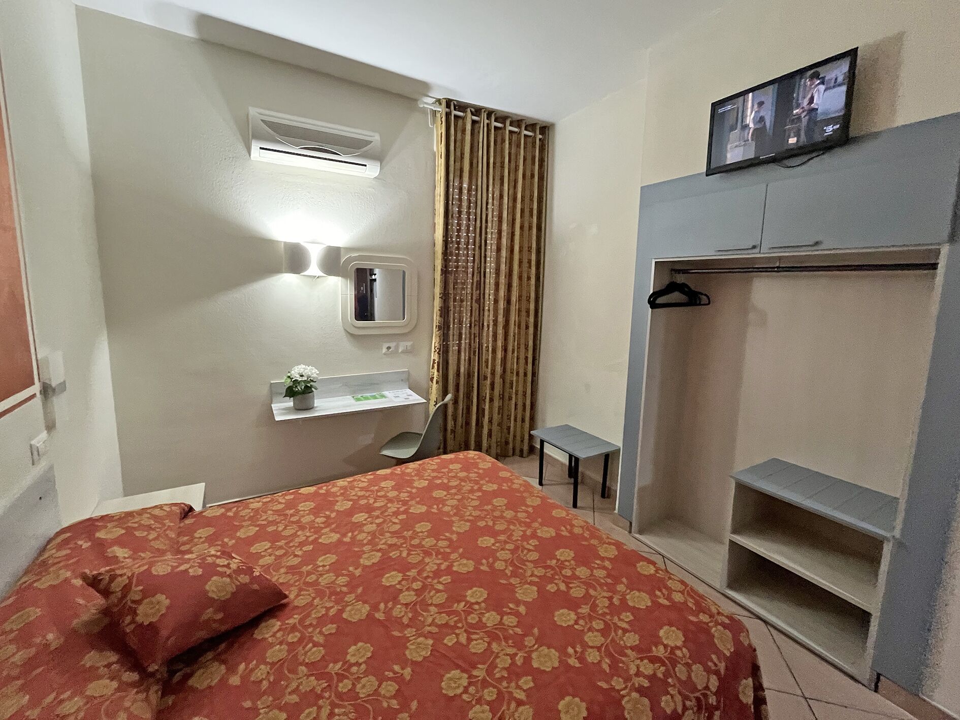 Ein Hotelzimmer mit einem Bett, einem kleinen Schreibtisch mit Pflanze, einem Spiegel, einem an der Wand befestigten Fernseher und einem Kleiderschrank.