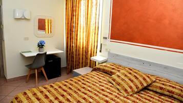 Quarto de hotel com cama, escrivaninha, cadeira, mesinha e janela com cortinas.