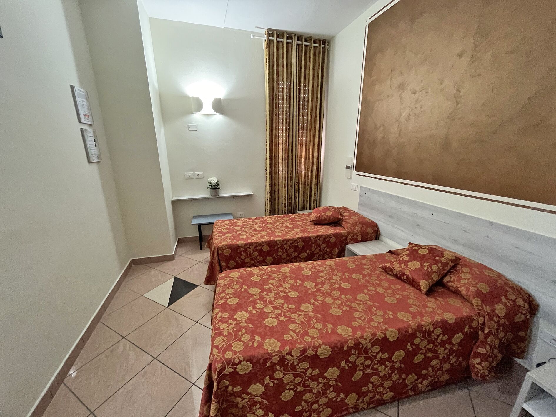 Ein Zimmer mit zwei Betten, einem Schreibtisch, einem Stuhl, einem kleinen Tisch mit Pflanze und einem Vorhang.