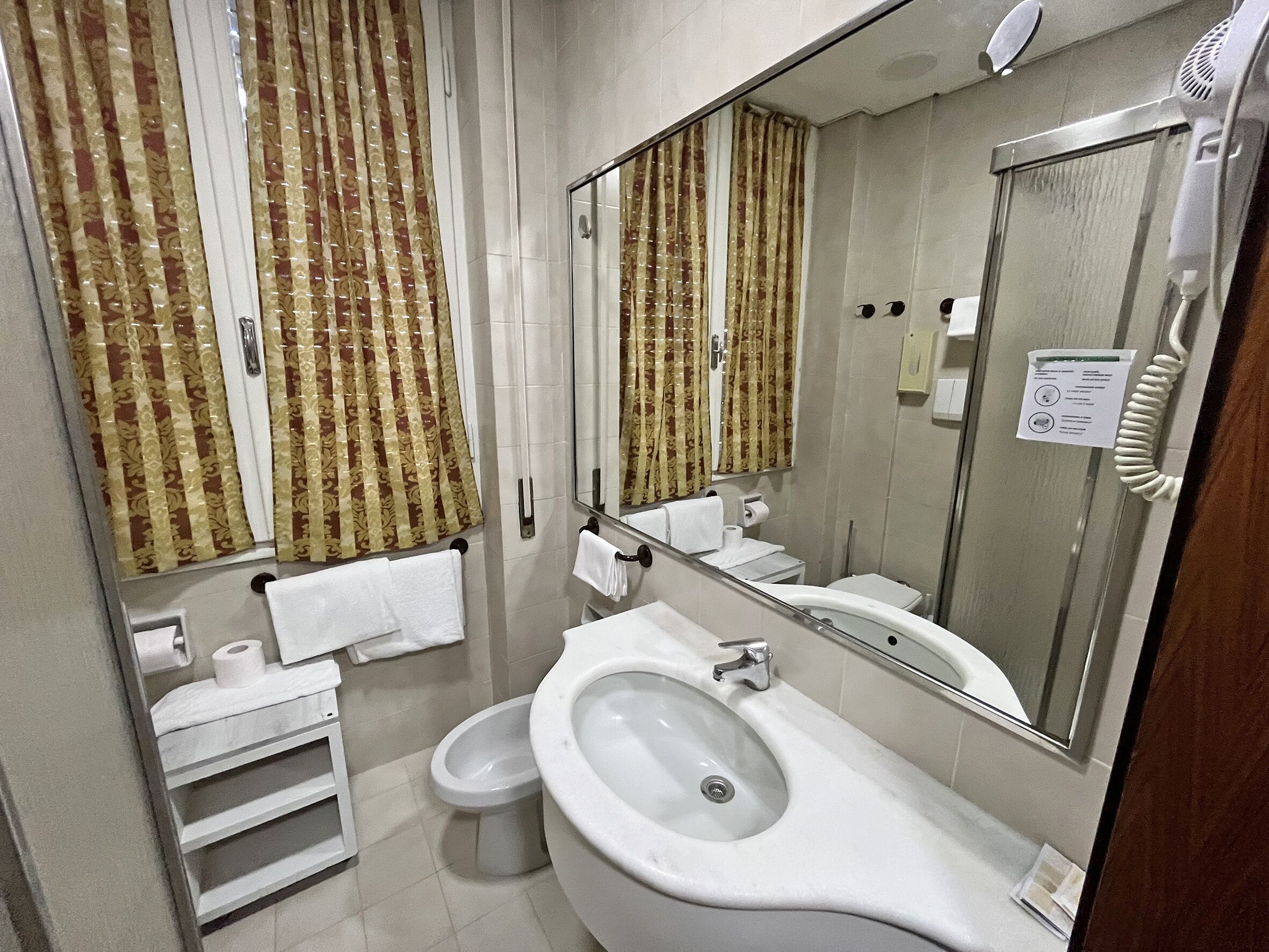 Un baño con lavamanos grande, inodoro y espejo. Hay toallas colgadas en la pared y un dispensador de servilletas de papel.