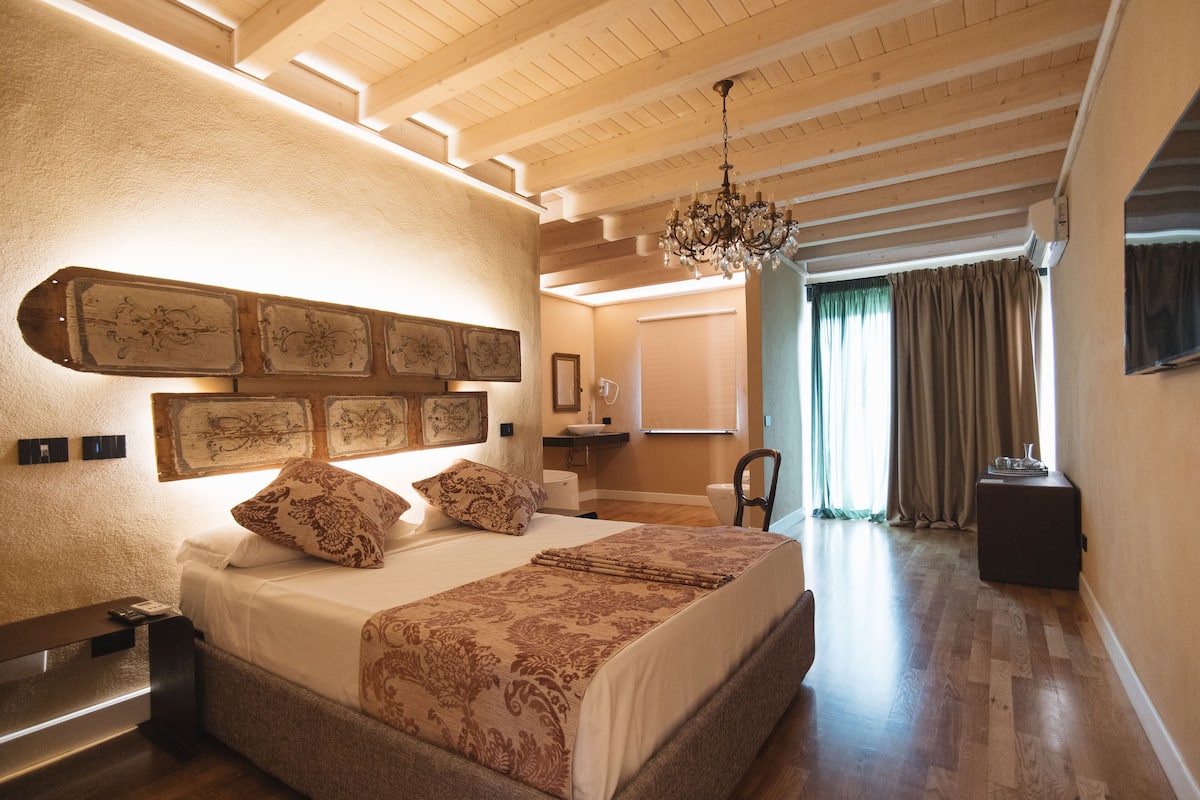 A bedroom with a large bed, a chandelier, a wooden ceiling, and a view of the bathroom.