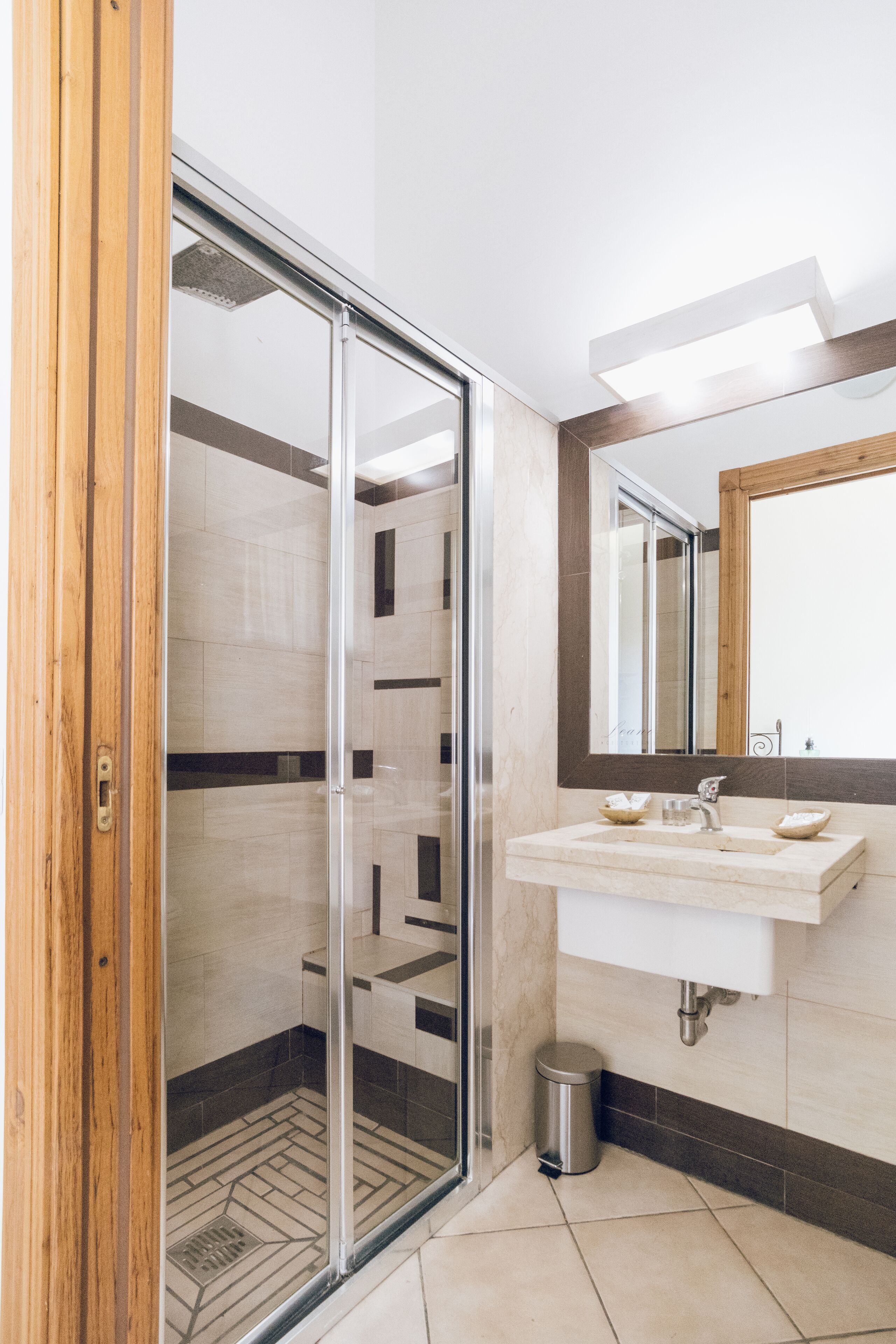 A modern bathroom with a glass shower enclosure, a wooden door, a white sink, and a mirror above it.