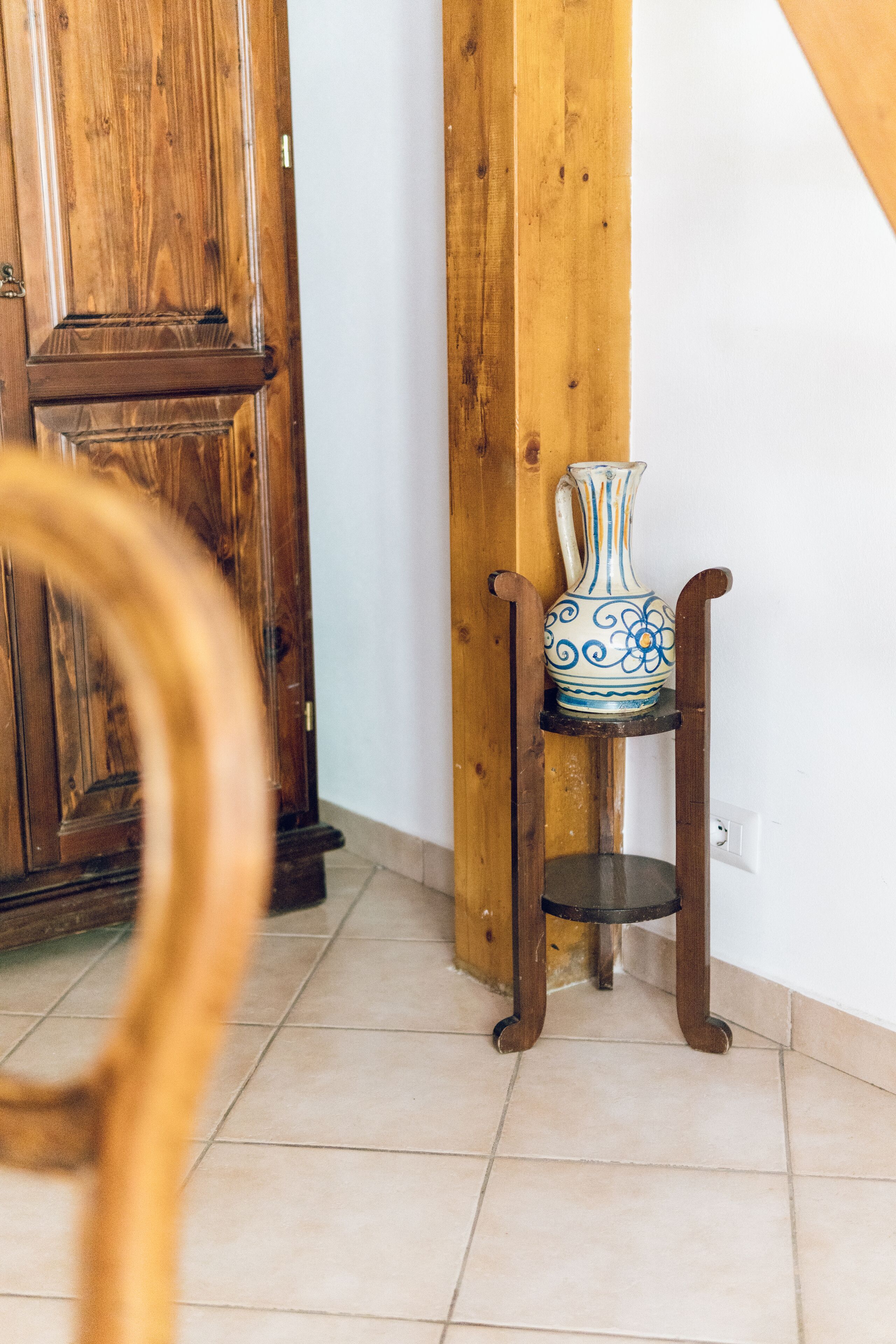 Een houten stoel met een vaas op een standaard in een kamer met een houten deur en een betegelde vloer.