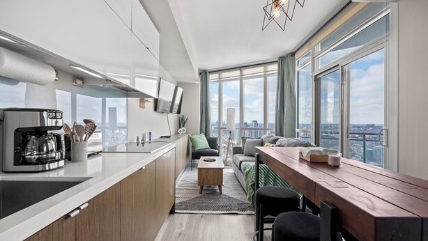 A modern kitchen with a coffee maker, a dining area with a wooden table and chairs, and a large window with a city view. - Elegant Downtown Apartments (Toronto)