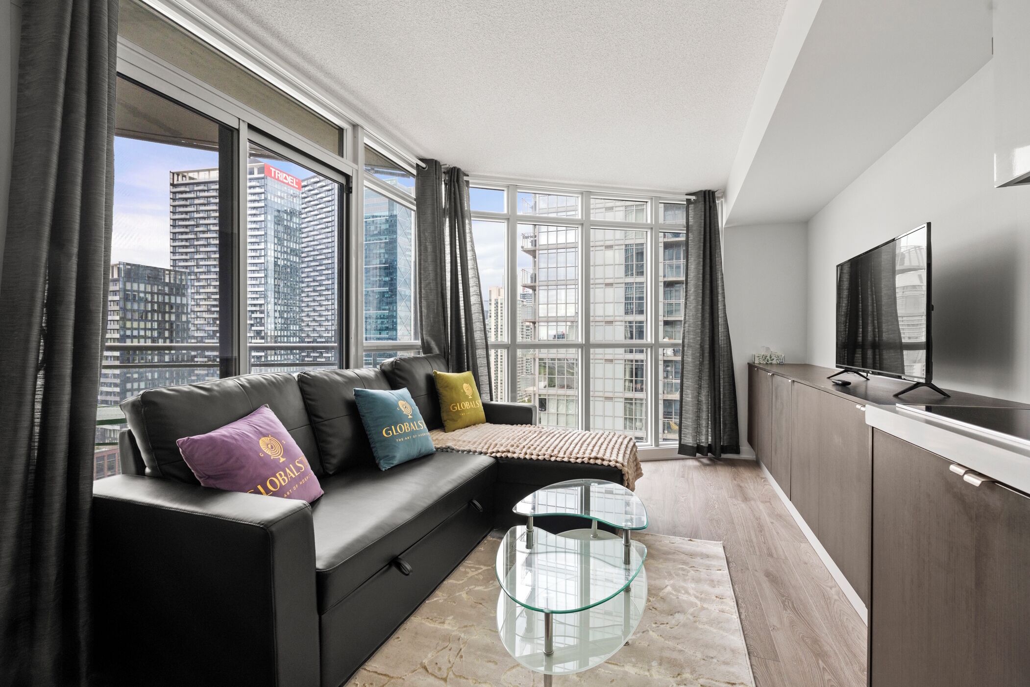 Un salon moderne avec un canapé, une table basse en verre et un téléviseur. De grandes fenêtres offrent une vue sur la ville.