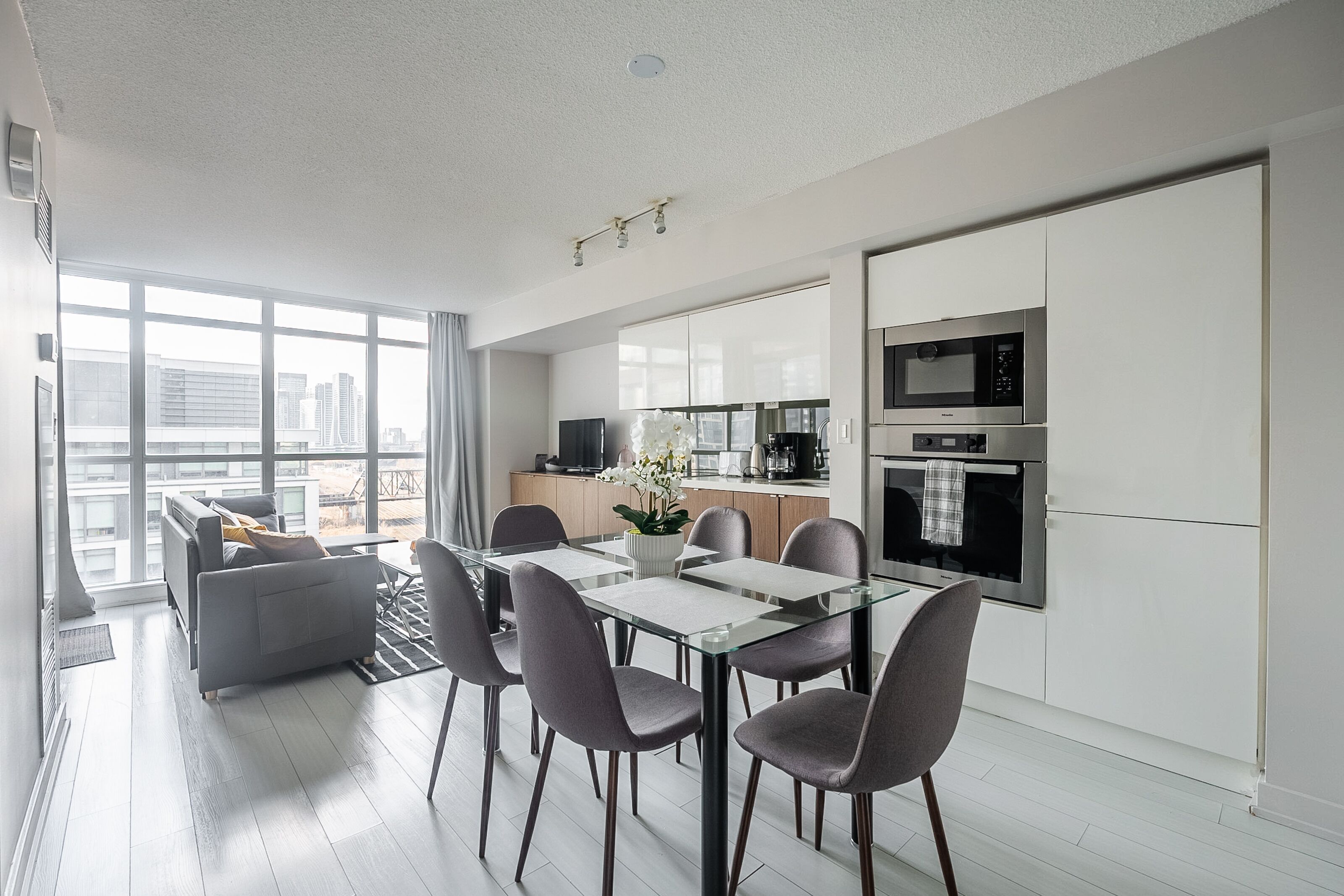 A modern kitchen with white cabinets, a dining area with a glass table, and a city view through large windows.