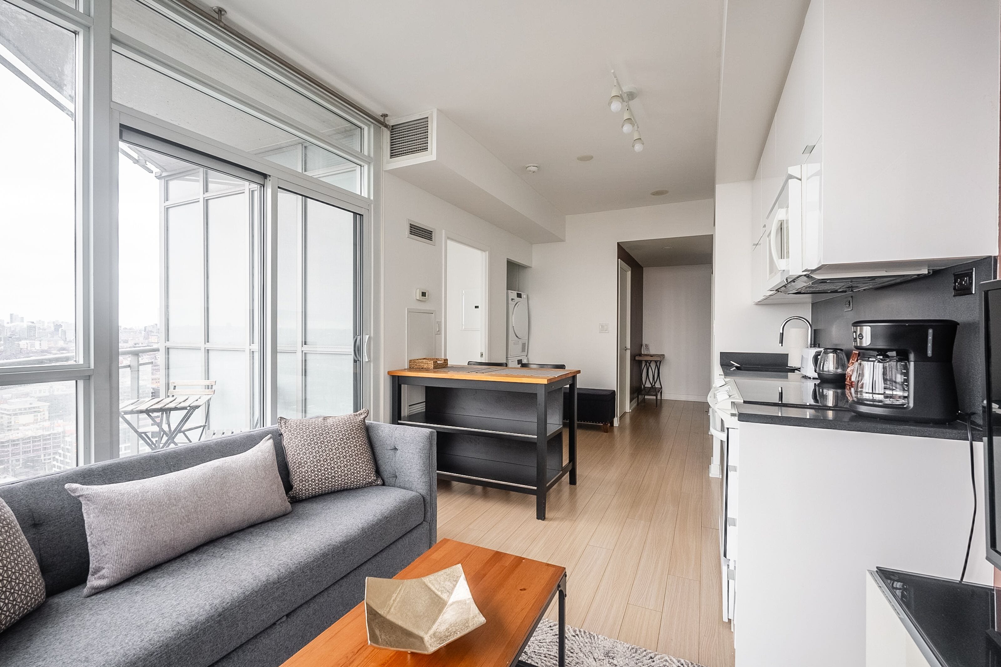 A modern apartment with a living area featuring a gray sofa, a wooden coffee table, and a large window offering a city view.