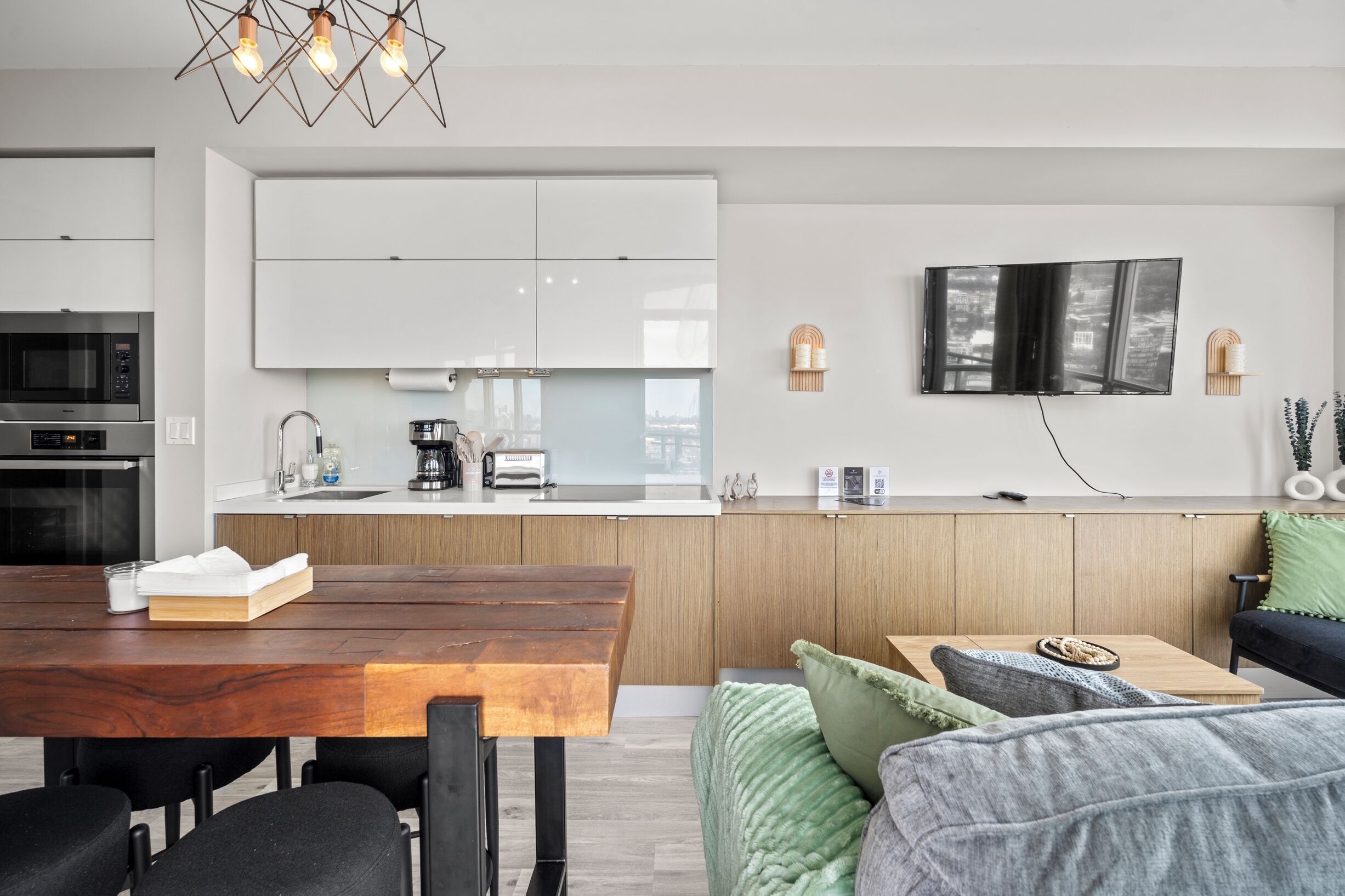 Een moderne keuken met witte kasten, een houten eettafel en een flatscreen tv aan de muur.
