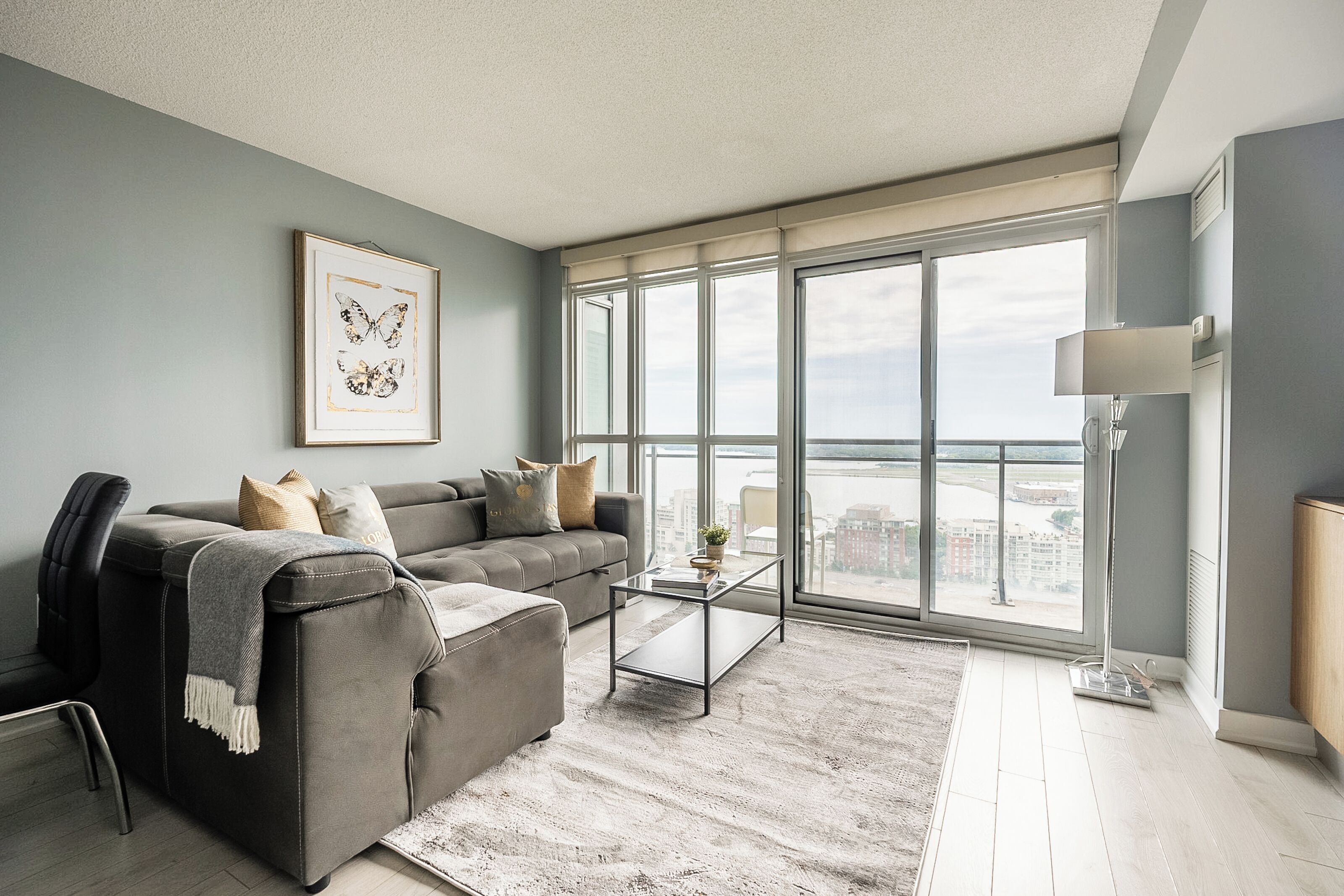 A modern living room with a grey sofa, a glass coffee table, and a large window offering a city view.