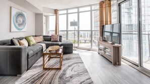 A modern living room with a sectional sofa, a coffee table, and a TV on a wooden stand. Large windows offer a view of the cityscape. - Elegant Downtown Apartments (Toronto)