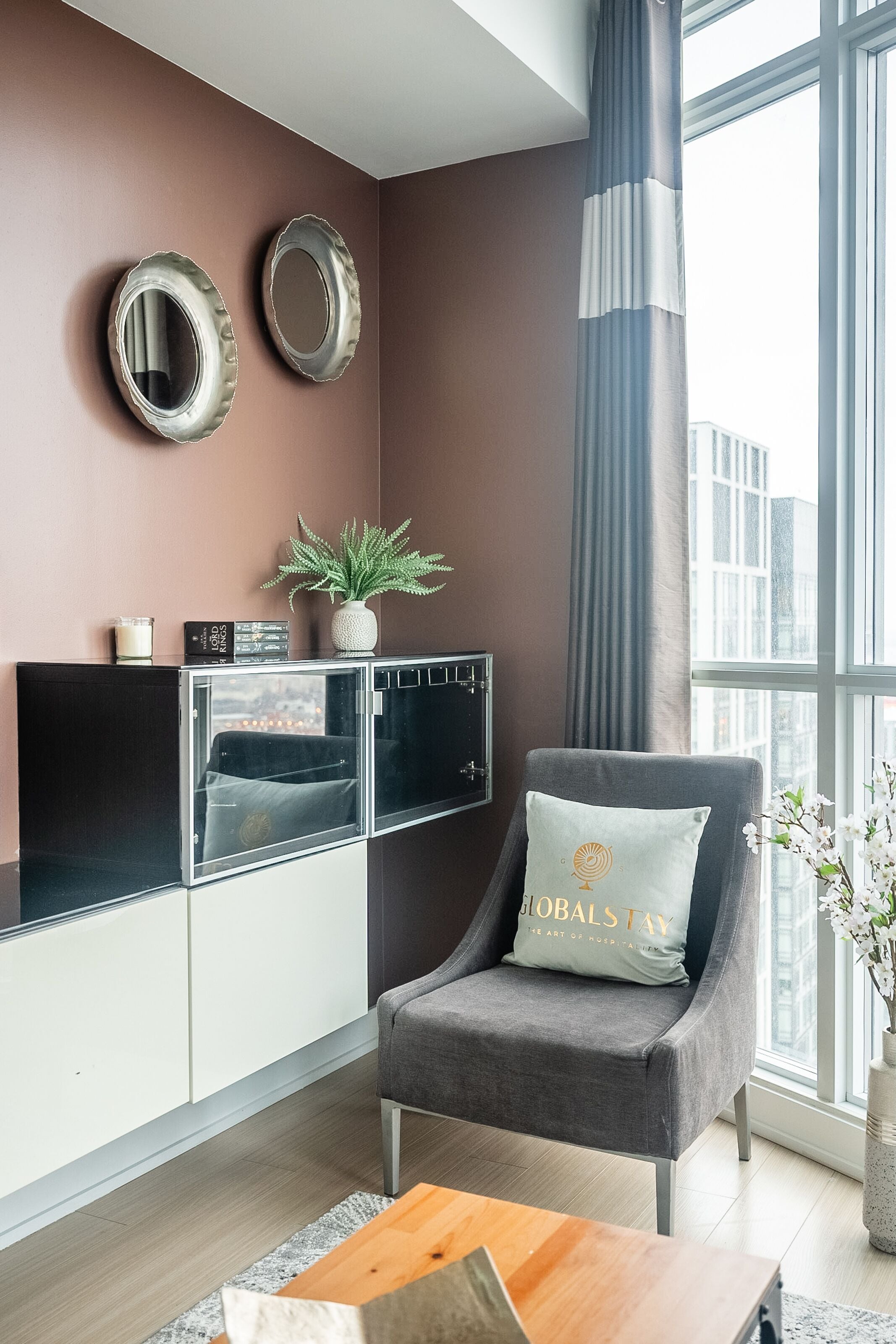 A modern living room with a grey armchair, a coffee table, a cabinet with glass doors, and a large window with a view of the city.