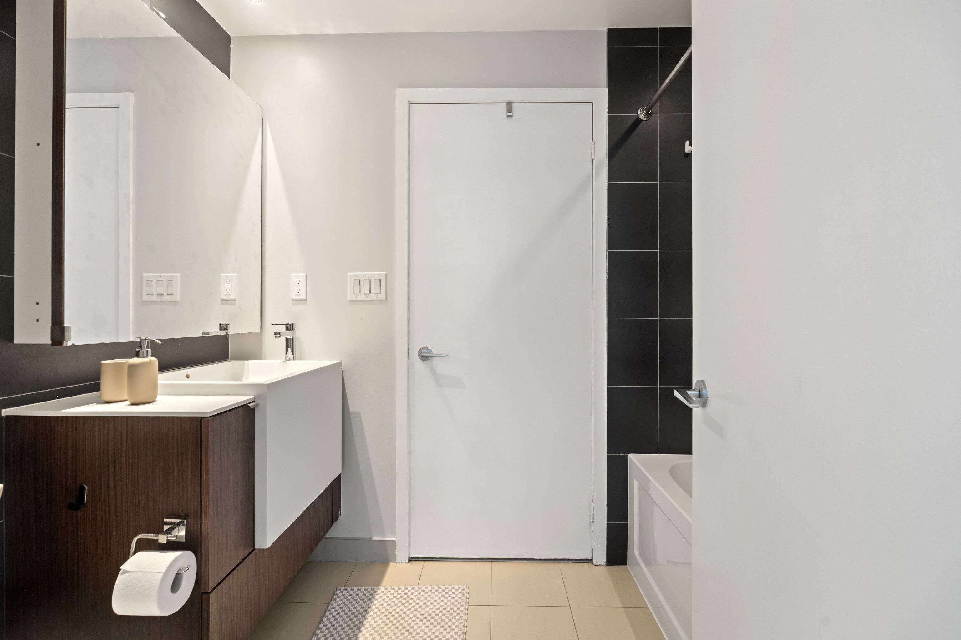 A modern bathroom with a white sink, dark tiles, a shower, and a door.