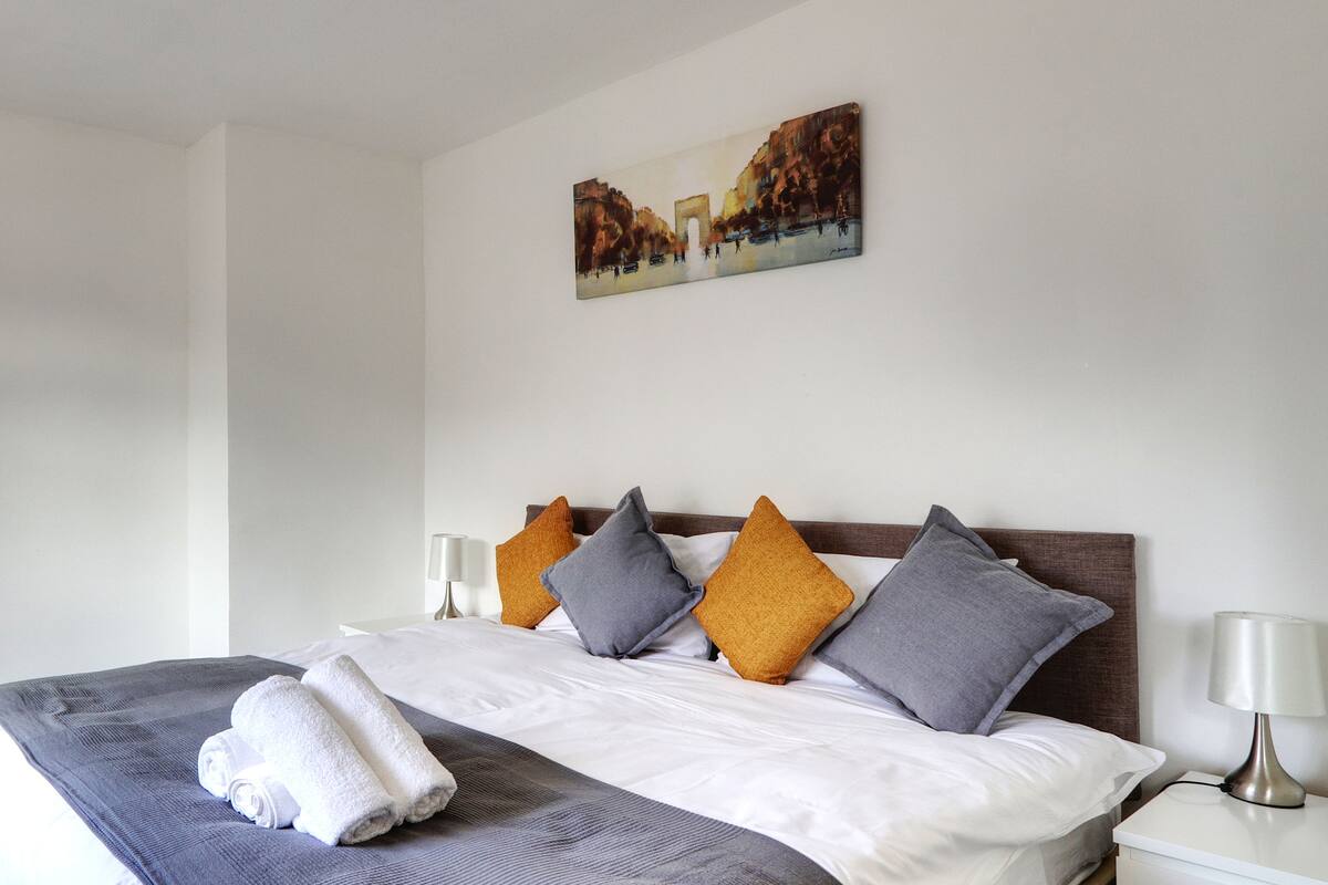 A neatly made bed with a blue and white bedspread, two white lamps on either side, and a painting above the bed.