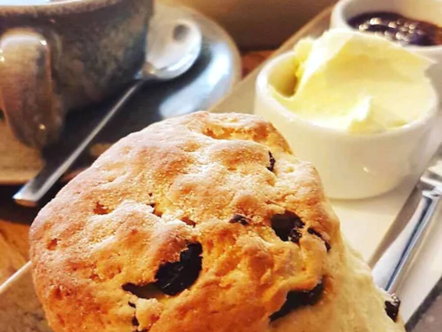 Un muffin à la croûte friable et contenant des morceaux de fruits visibles, servi avec un petit ramequin de beurre et une tasse de café sur un plateau.