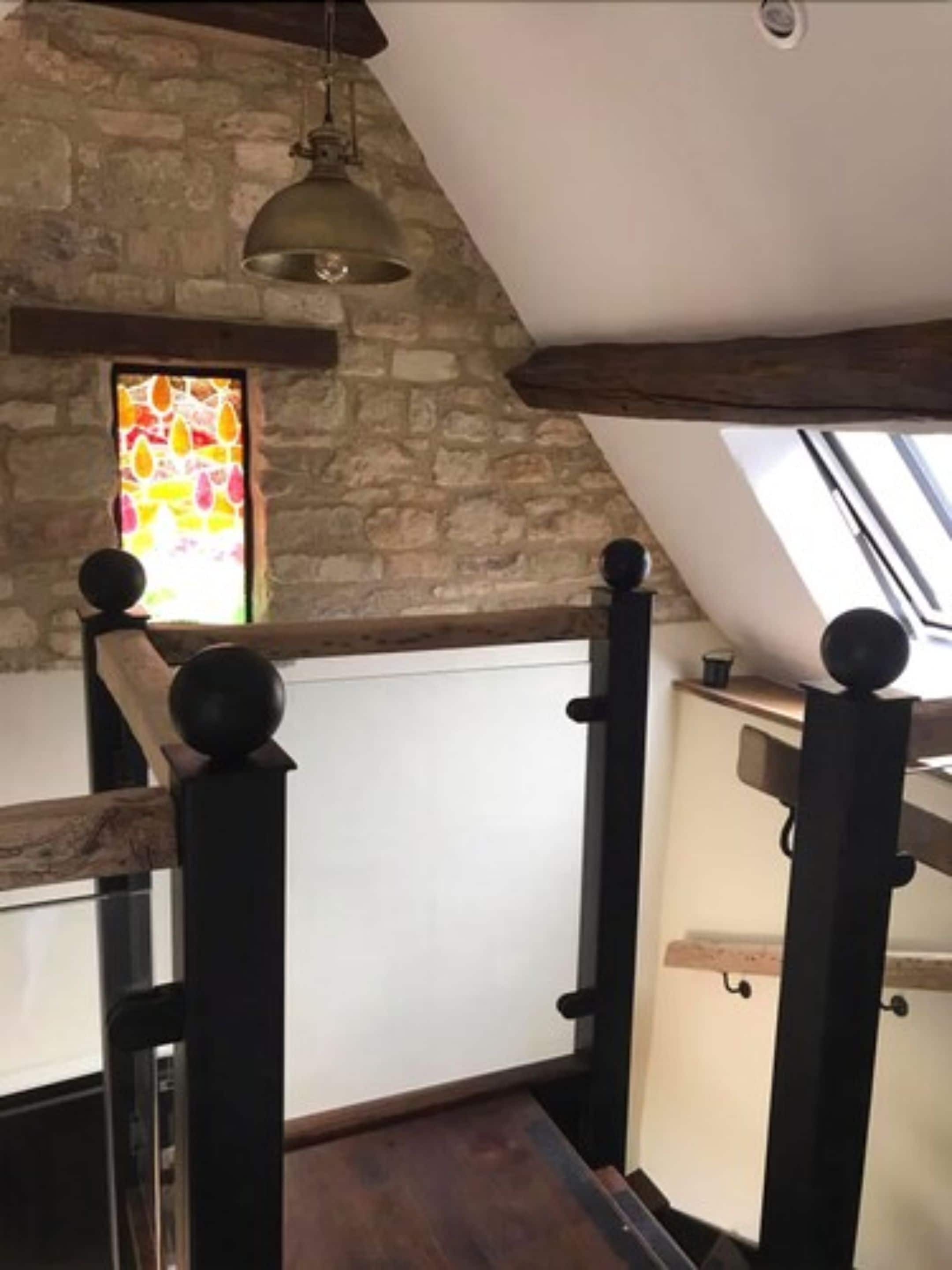A room with a sloped ceiling, stone walls, a wooden staircase with black balusters, and a stained glass window.