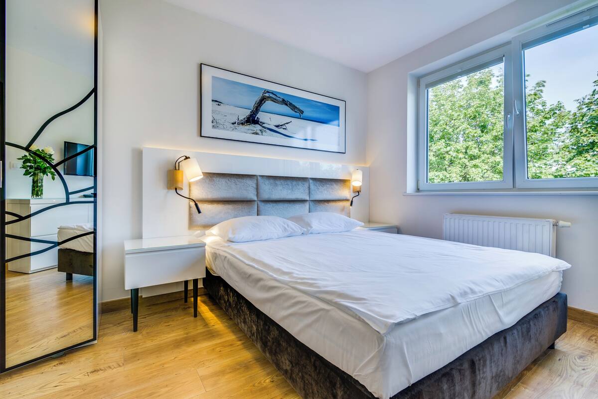 A modern bedroom with a large bed, bedside tables, a mirror, and a window with a view of trees.