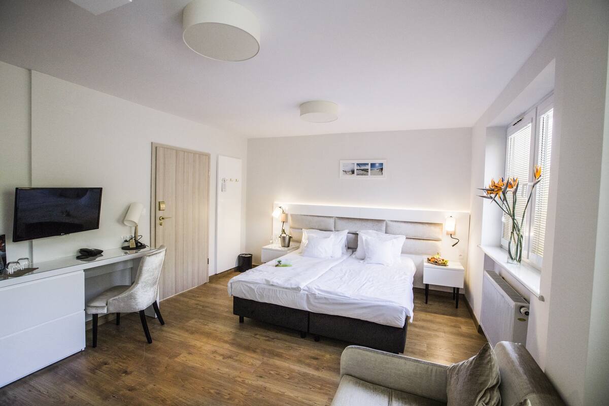 A hotel room with a bed, bedside tables, a desk with a chair, a television, and a window with flowers.