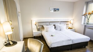 A hotel room with a double bed, bedside tables, a desk with a chair, a lamp, and a window with flowers.