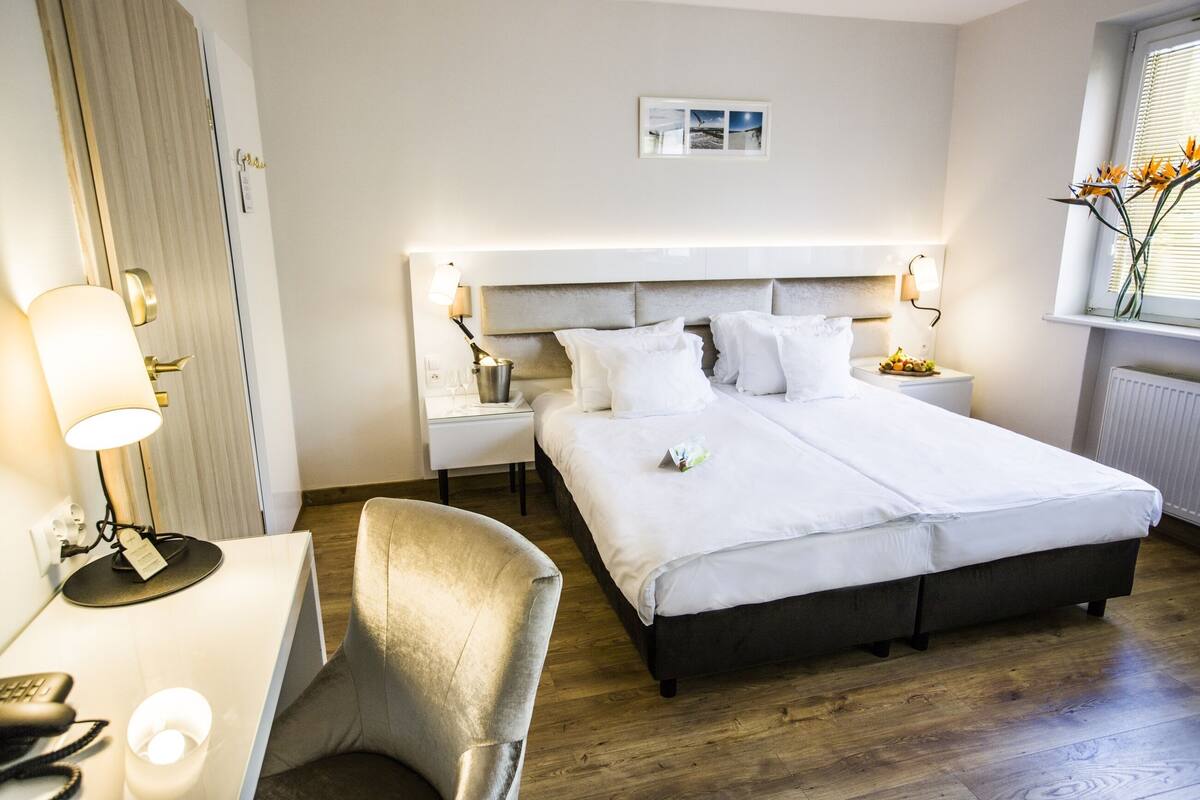 A hotel room with a double bed, bedside tables, a desk with a chair, a lamp, and a window with flowers.
