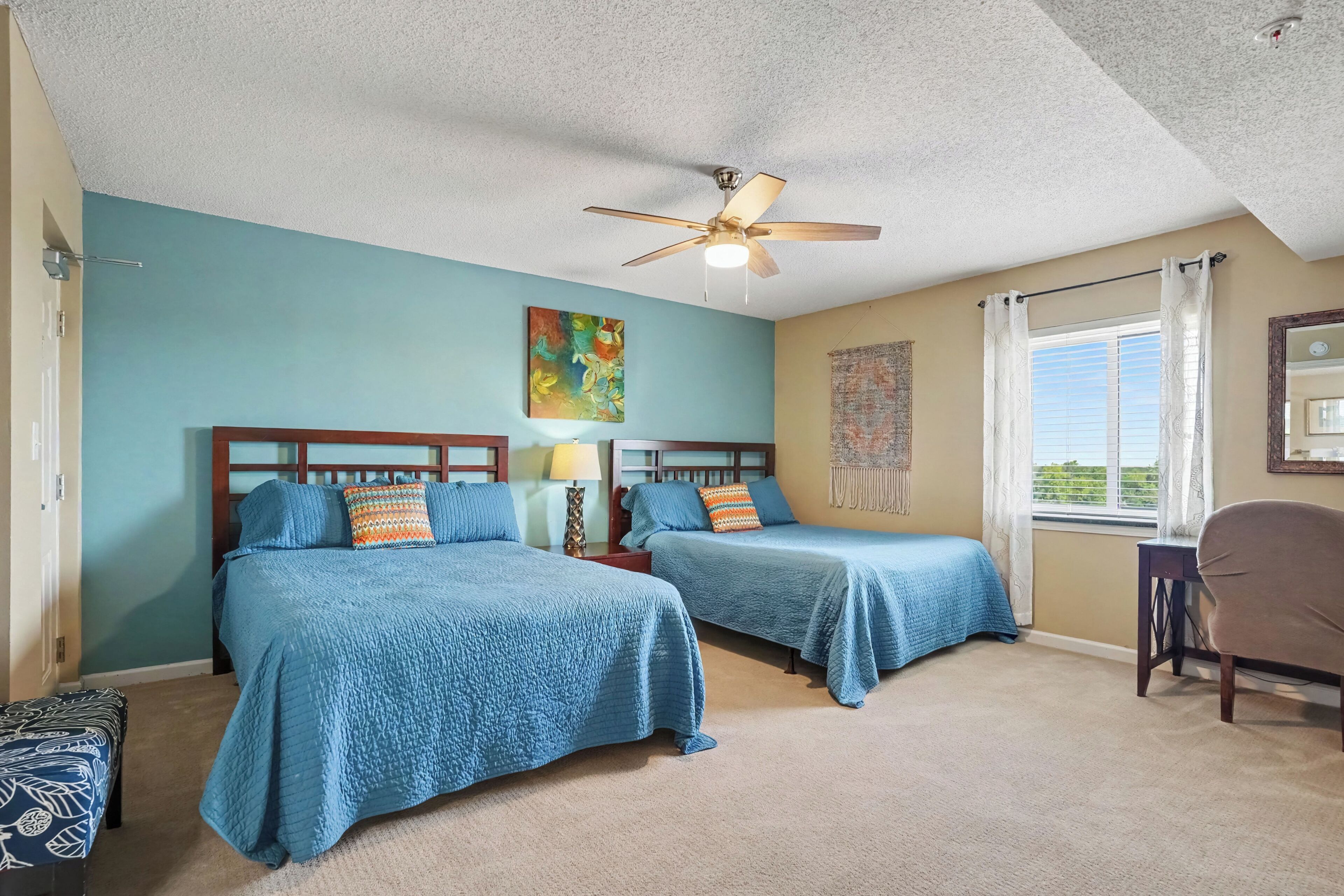 A hotel room with two beds, a ceiling fan, a painting, and a window with curtains.