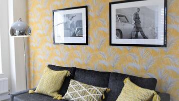 Una stanza con un divano grigio scuro, carta da parati gialla con motivi decorativi e due fotografie in bianco e nero appese al muro.