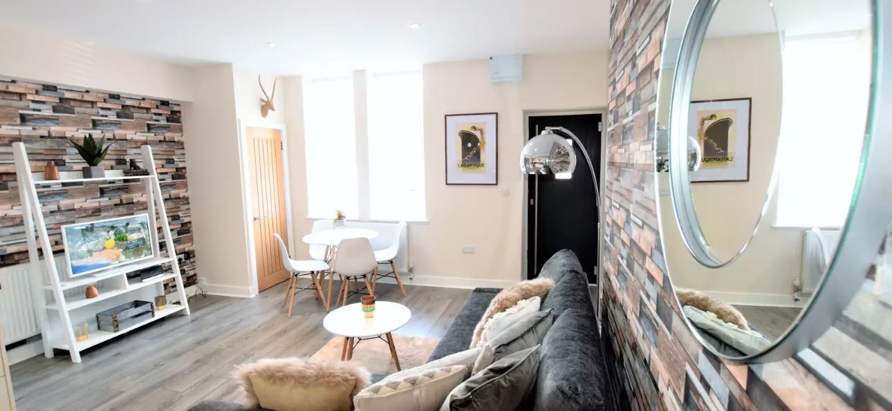 Un soggiorno moderno con divano, uno specchio rotondo, una TV su un mobile bianco, un'area pranzo con tavolo e sedie e un muro con decorazione a mattoni.