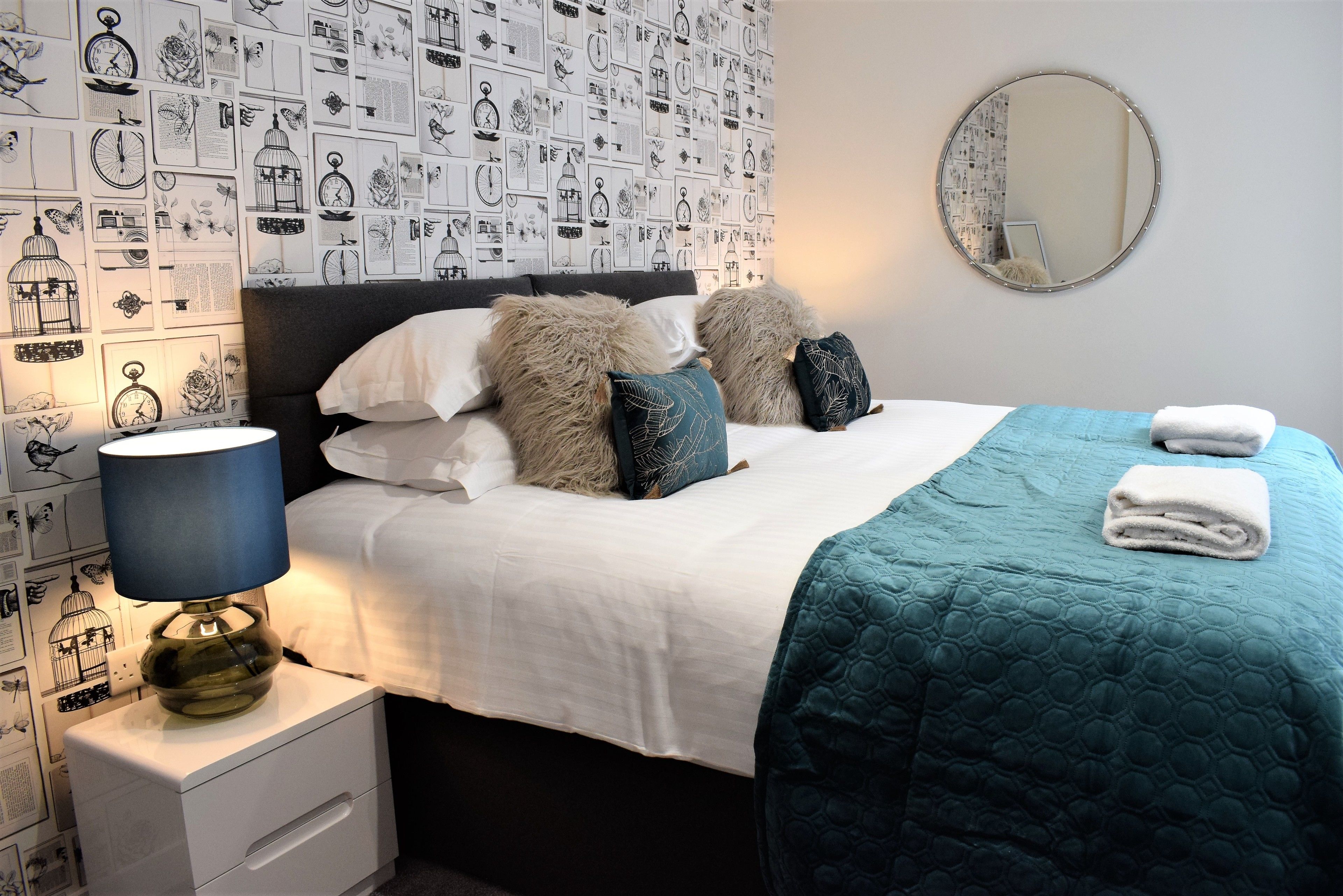 A neatly made bed with a teal quilt, fluffy pillows, and a white comforter. A round mirror hangs on the wall above the bed.